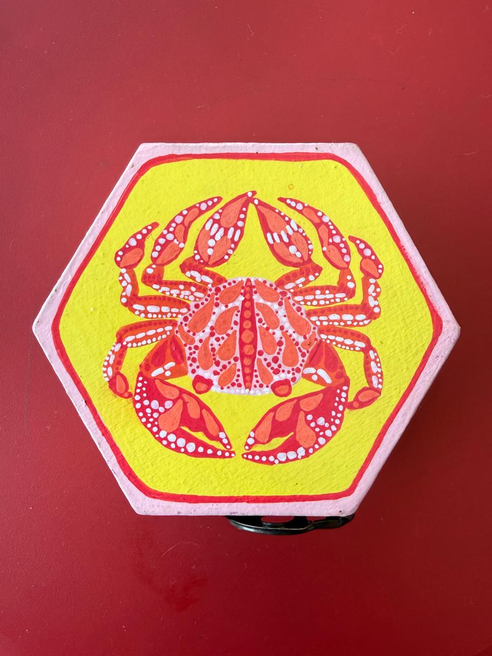 A small hexagonal trinket box with a red crab painted on a yellow ground.