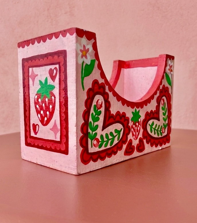 A pink napkin holder with a strawberry side panel and heart-and-leaf front.